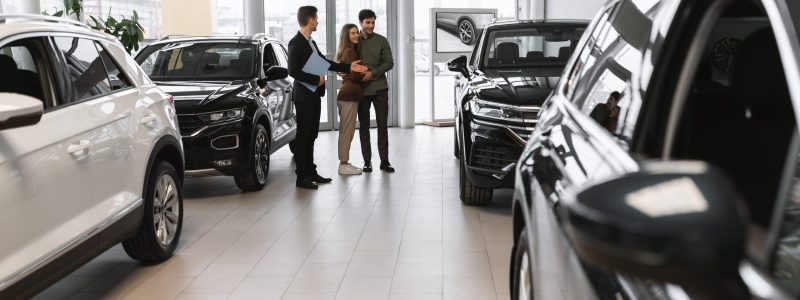 Full length of millennial couple choosing new car, having discussion with auto salesman at vehicle dealership. Cheerful young Caucasian family selecting automobile, communicating with showroom manager
